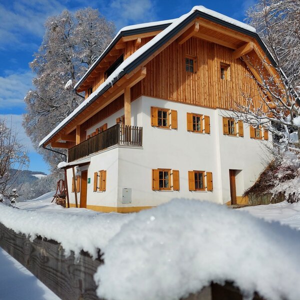 The Alpine Hut's exterior in winter, featuring wooden upper levels, white lower walls, a balcony, and snow-covered surroundings.