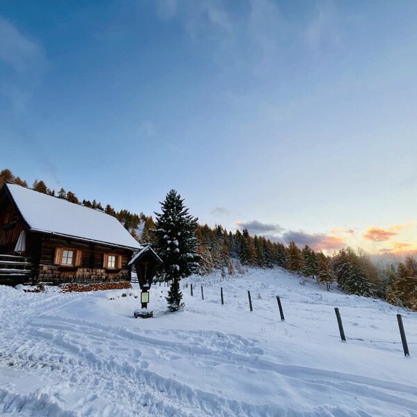 The snow-covered Alm with lit windows and stacked firewood, surrounded by a winter landscape.