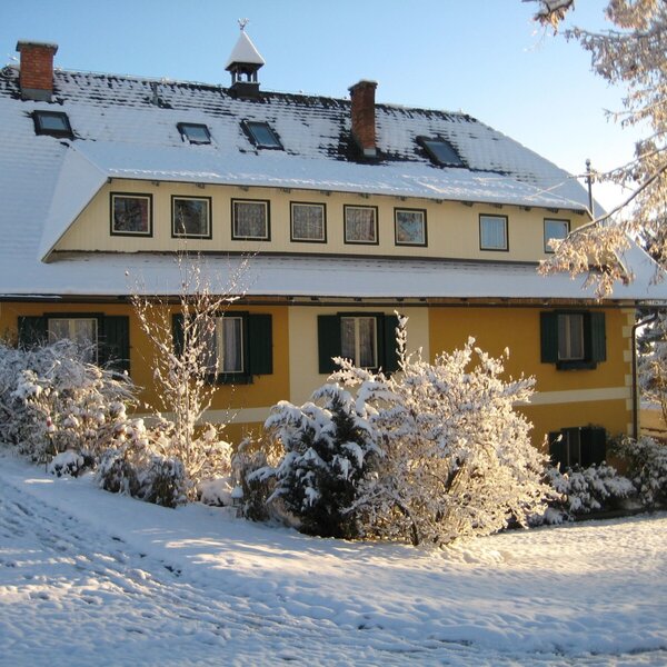 Die Außenansicht des Bauernhofs im Winter, mit gelber Fassade, grünen Fensterläden und schneebedecktem Dach.