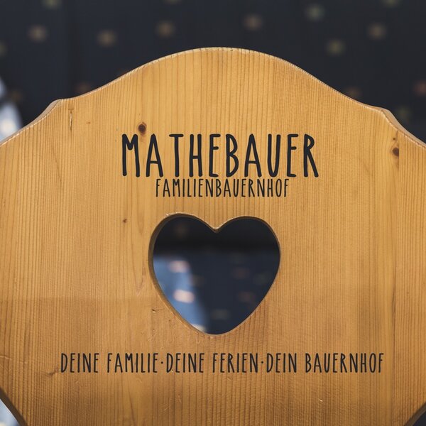A wooden chair backrest at the Farm House features the name 'Mathebauer Familienbauernhof', a heart-shaped cutout, and the phrase 'Your Family Your Holidays Your Farmhouse'.