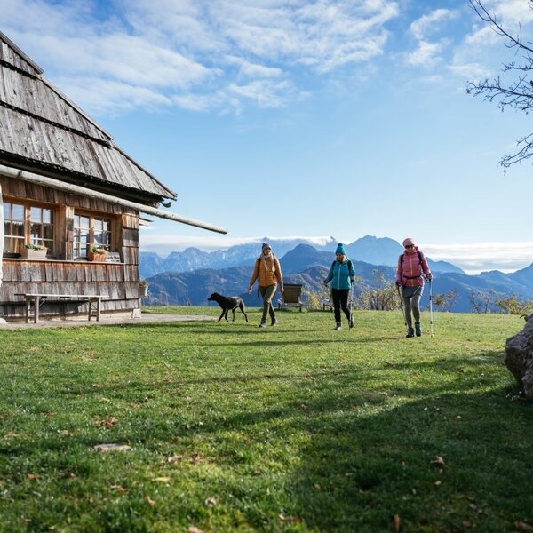 Das Gebäude des Hofs mit einer Gruppe Wanderer und einem Hund auf der Wiese und Blick auf die Berge.