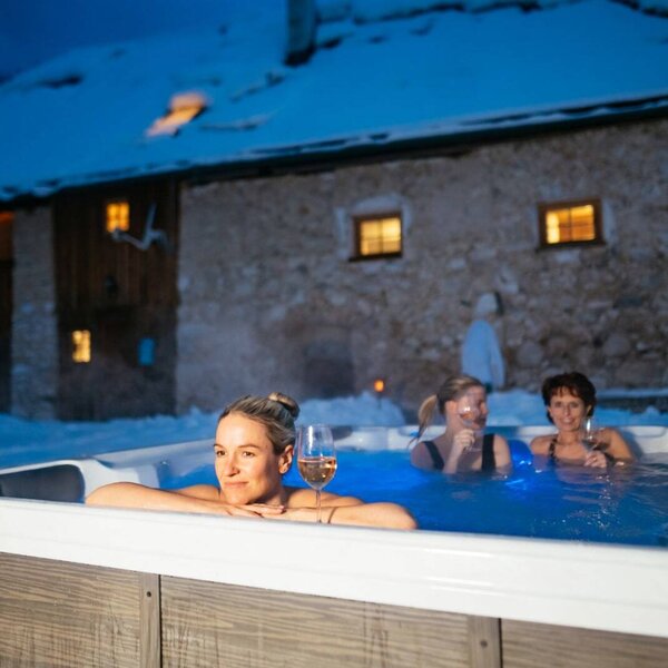 The Alpine Hut offers an outdoor hot tub for guests, set in the snow at night, with the illuminated building visible behind it.