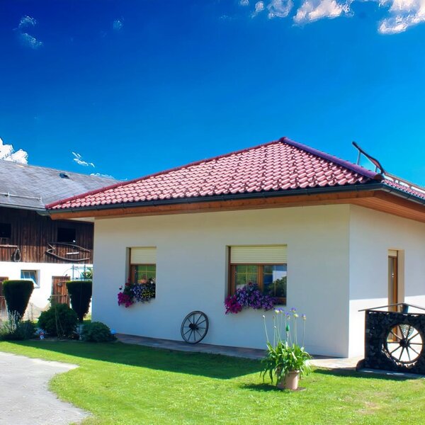 Der Bauernhof mit dem weiß verputzten Hauptgebäude, rotem Ziegeldach, der angrenzenden Scheune und einem Rasenbereich mit Terrasse.