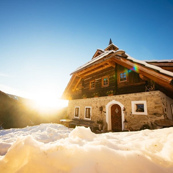 Die Alm in traditioneller Holz- und Steinbauweise in einer verschneiten Berglandschaft bei Wintersonne.