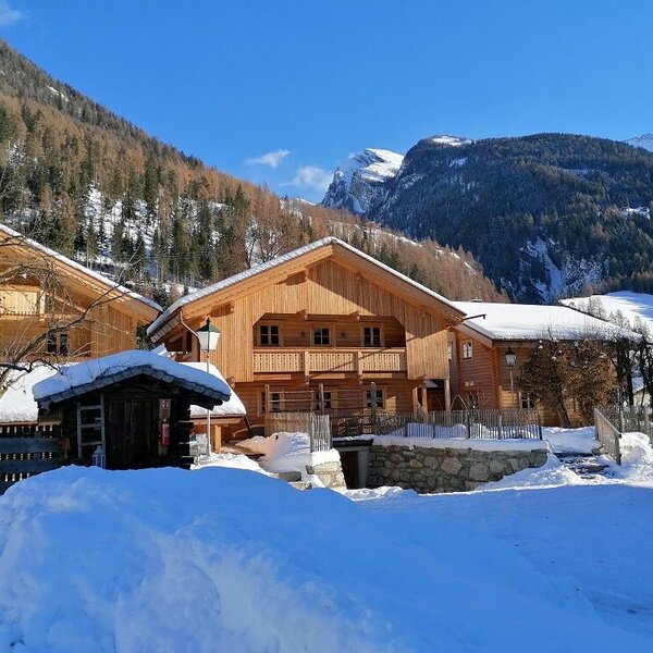 Der winterliche Hof mit seinen Holzhäusern und dem Traktor, vor der Kulisse schneebedeckter Berge.