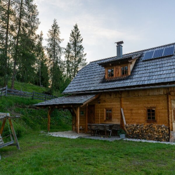 The Alpine Hut, featuring a wooden exterior, shingle roof with solar panels, a covered outdoor dining area, and a wooden swing in the garden.