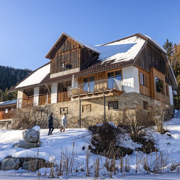 Das Ferienhaus mit Balkon, eingebettet in eine verschneite Winterlandschaft mit angrenzendem Wald und gefrorenem Wasser im Vordergrund.