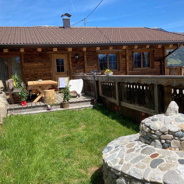 Chalet mit einer Holzterrasse, ausgestattet mit Tisch und Stühlen, umgeben von einer grünen Wiese und Bergblick.