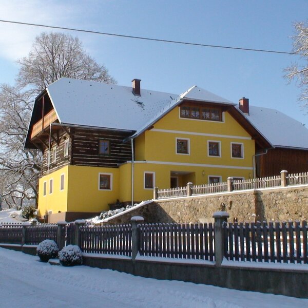 The farmhouse exterior during winter, featuring a yellow lower facade, a wooden upper level, and a snow-covered roof.
