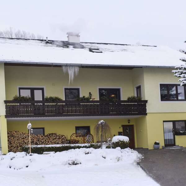 The yellow Farm House exterior in winter, featuring a snow-covered roof, icicles, smoke from the chimney, a wooden balcony, and a stack of firewood on the ground floor.