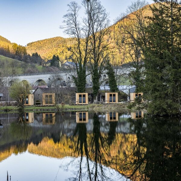 Die kleinen Holzhäuser des Bauernhofs am Wasser, mit Reflexionen der Umgebung und Blick auf Berge und eine Brücke.