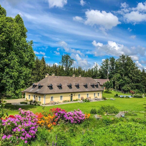 Der Bauernhof mit gelber Fassade und braunem Dach, umgeben von einem weitläufigen Garten mit blühenden Büschen, Pool und angrenzendem Wald.