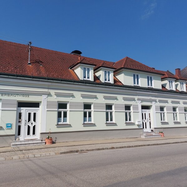 Die Außenansicht des Bauernhofs mit heller Fassade, zwei Eingängen, einem Ziegeldach und Gaubenfenstern, erbaut im Jahr 1922.