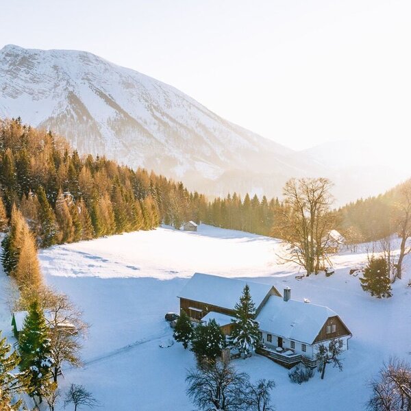 The snow-covered farmhouse is situated in a sunny winter landscape with surrounding forests and mountains.