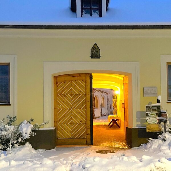 The entrance to the farmhouse features an open wooden door with a diamond pattern, leading to a brightly lit interior, surrounded by snow.