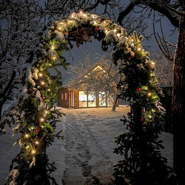Ein verschneiter Bogen mit Lichterketten und roten Kugeln führt zum beleuchteten Chalet im Obstgarten des Bauernhofs.