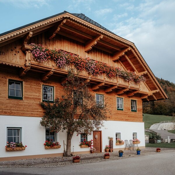 The traditional Farm House exterior features a wooden upper floor with flower-filled balconies, a white lower level, and an entrance with outdoor seating.
