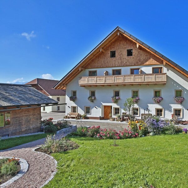 The main building of the farmhouse, featuring a wooden balcony, flower boxes, and a front garden with a lawn and gravel path.
