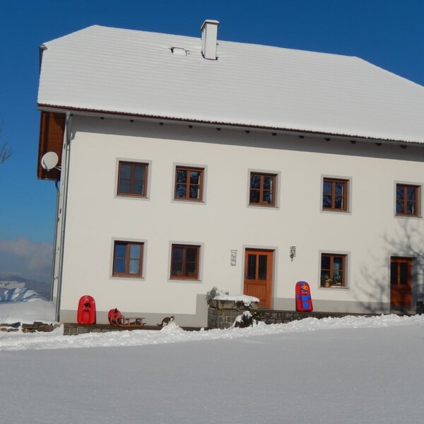 The white farmhouse exterior, featuring a snow-covered roof and ground, with red sleds positioned near the entrance.