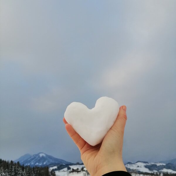 A hand holding a heart-shaped snowball in the snowy mountain landscape surrounding the farmhouse.