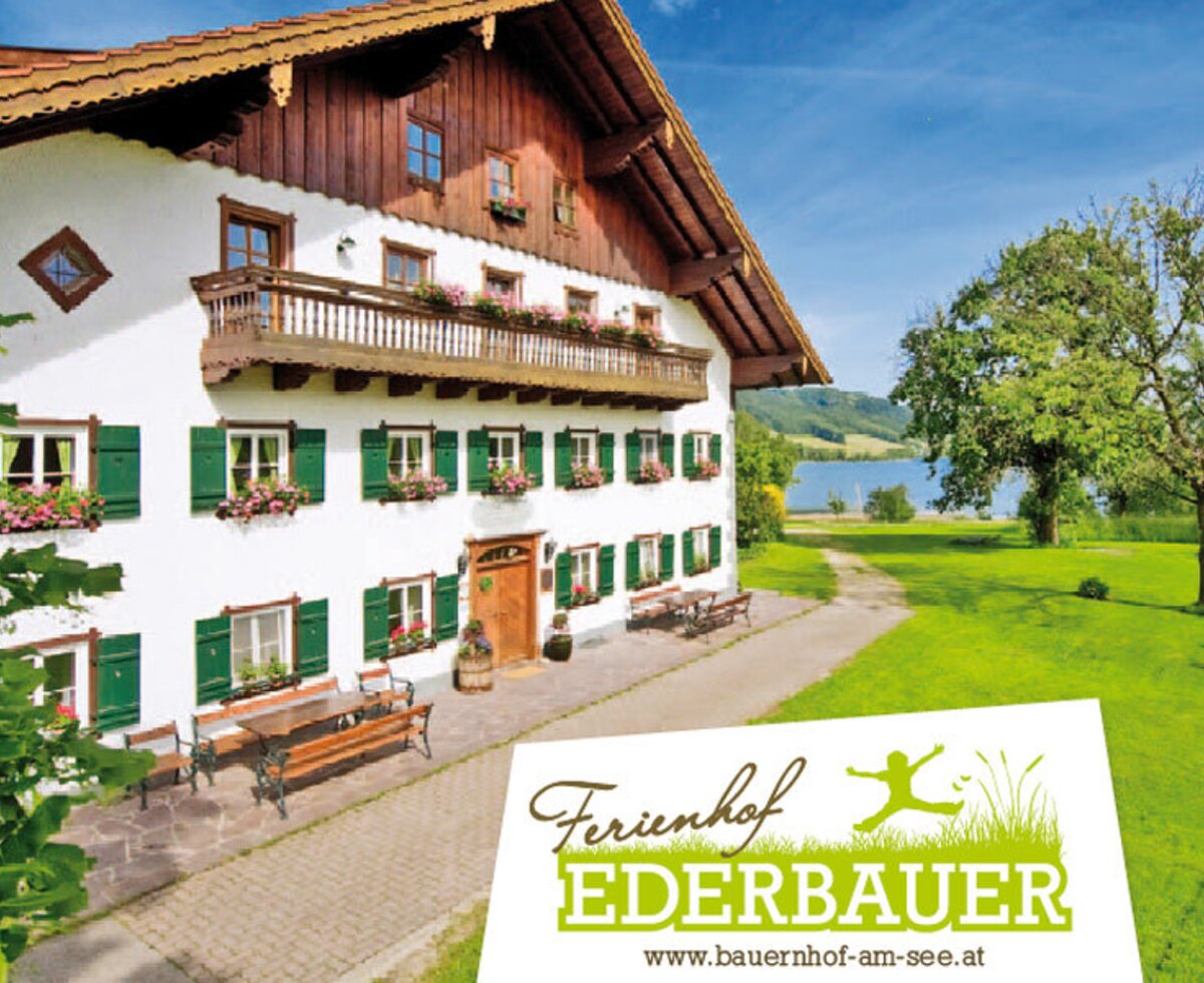 Außenansicht des Bauernhofs mit weißer Fassade, grünen Fensterläden, einem Balkon und Blumenkästen, dazu ein Sitzbereich im Freien mit See- und Bergblick.