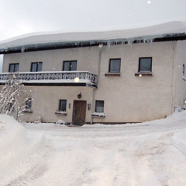 The farm house exterior in winter, featuring a snow-covered roof with icicles, a balcony, and a snowy driveway.