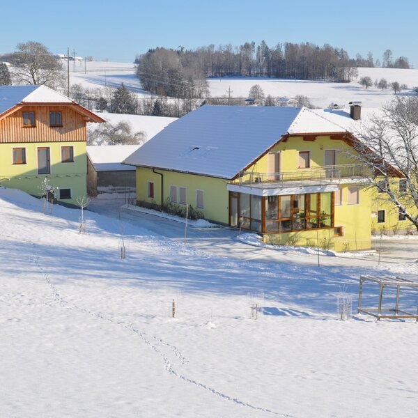 Die Gebäude des Bauernhofs im Winter, mit dem Wintergarten und einem Schaukelgestell auf dem verschneiten Spielplatz.