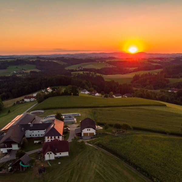 Aerial view of the farmhouse complex amidst green fields and distant hills, illuminated by a sunset.