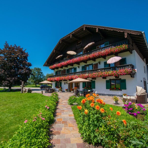 The Farm House exterior offers balconies with flower boxes, green shutters, and a ground-floor patio with outdoor seating and umbrellas, surrounded by a landscaped garden and green lawns.