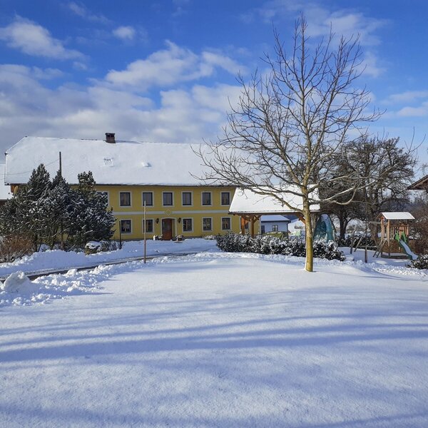 Die schneebedeckte Außenansicht des Bauernhofs mit dem gelben Hauptgebäude und einem Kinderspielplatz.