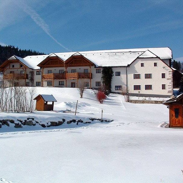 Der Bauernhof ist ein weißes Gebäude mit Balkonen und Fenstern, eingebettet in eine schneebedeckte Winterlandschaft mit Bäumen im Hintergrund.