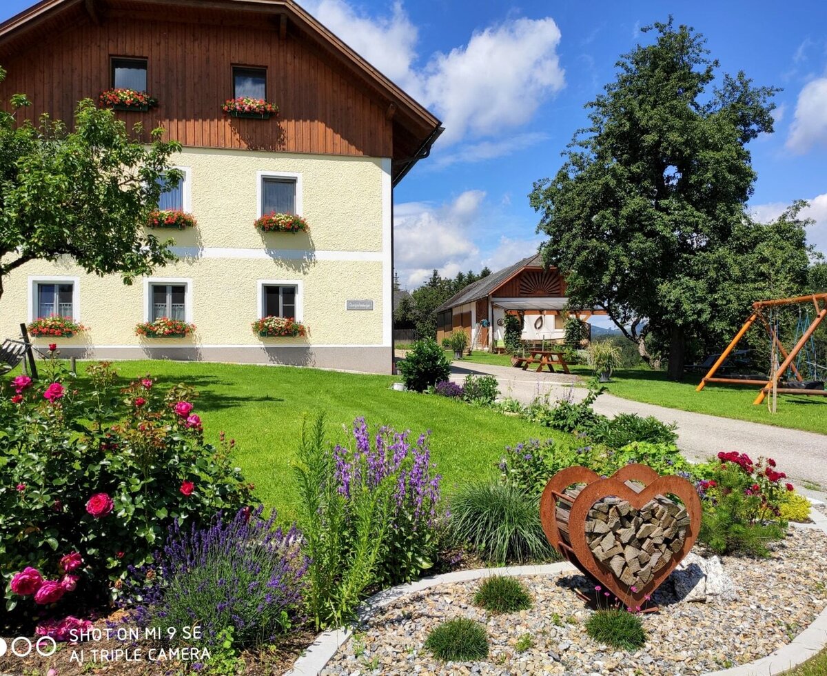 Der Bauernhof mit gelber Fassade und Blumenkästen, umgeben von einem Garten mit Rasenfläche, Blumenbeeten und einer Schaukel.