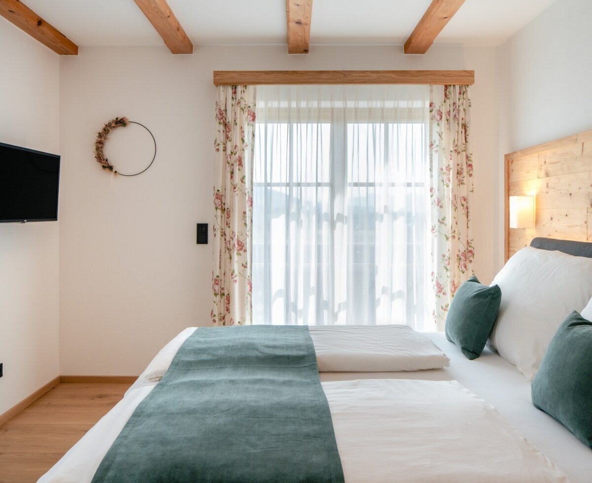 Schlafzimmer im Bauernhof mit Doppelbett, Wandfernseher, Holzbalken an der Decke, Holzboden und einem Fenster mit floralen Vorhängen.