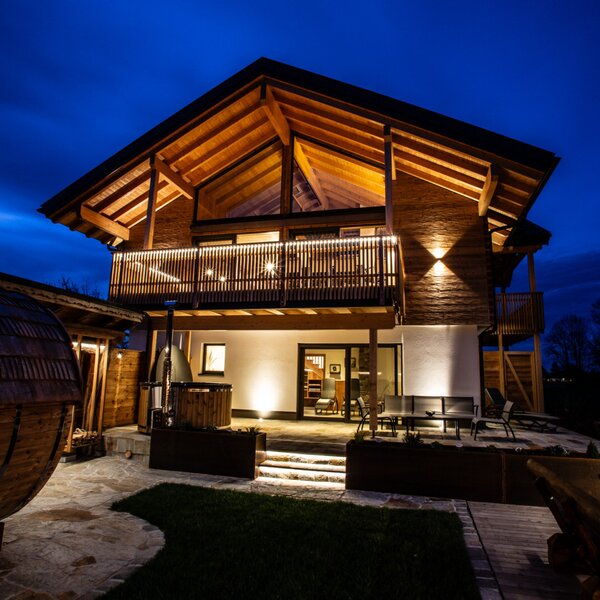 The illuminated Farm House at night, featuring wooden architecture, a balcony, and a ground-floor patio with outdoor seating, a hot tub, a pizza oven, and a barrel sauna.