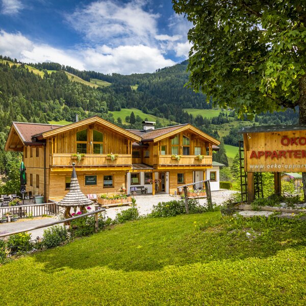 The wooden Farm House exterior, featuring balconies and a sign for Öko Appartements, set against a backdrop of green mountains and forests.