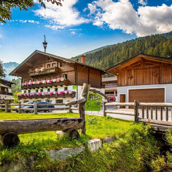 Der Bauernhof mit traditioneller Holzfassade, blumengeschmückten Balkonen und einem Nebengebäude, umgeben von grünen Wiesen und Bergen.