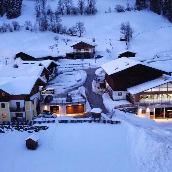 Der Bauernhof in seiner verschneiten Winterlandschaft, bestehend aus mehreren Gebäuden mit beleuchteten Fenstern.