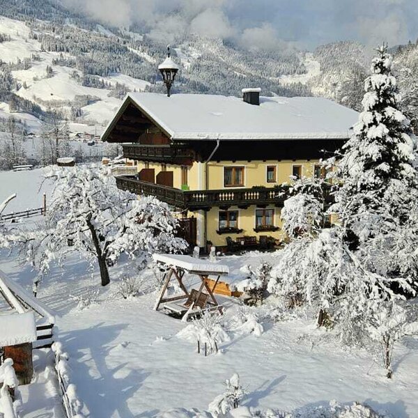 Der Bauernhof im Winter, mit Schnee bedecktem Garten inklusive Schaukel und einer Bergkulisse.