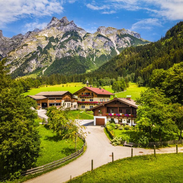 Außenansicht des Bauernhofs mit den umliegenden grünen Wiesen und den Bergen im Hintergrund.