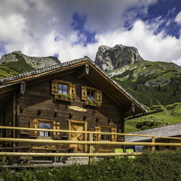 The wooden farmhouse exterior features window shutters, flower boxes, and a fenced terrace with views of the surrounding green mountains.