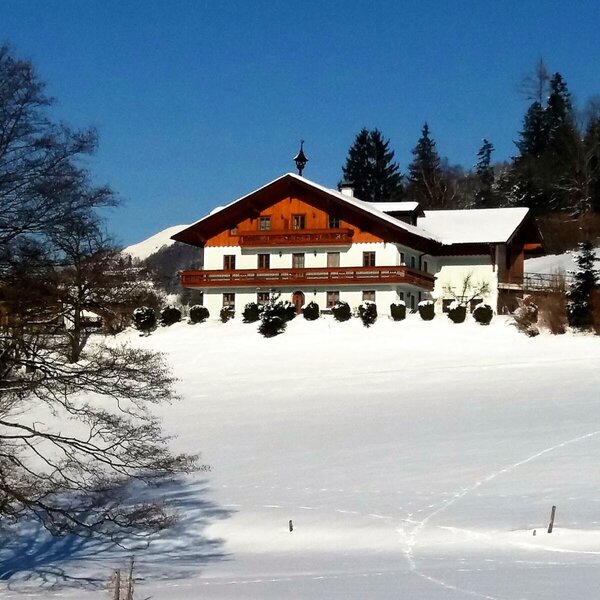Außenansicht des Bauernhofs im Winter, umgeben von einer schneebedeckten Landschaft und Bäumen unter blauem Himmel.