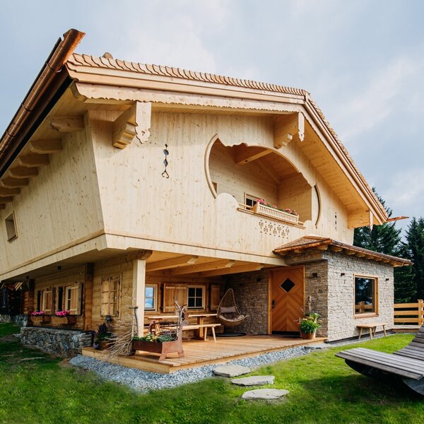 The exterior of the farmhouse features intricate wooden craftsmanship, a covered deck with outdoor seating, and a grass lawn with a wooden lounger.