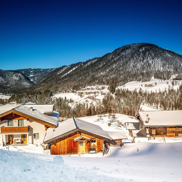The farm house and its outbuildings are set in a snow-covered mountain landscape under a clear sky.