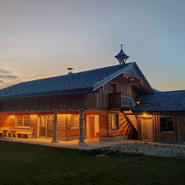 The Farm House exterior features a wooden construction, an illuminated covered terrace with outdoor seating, and a balcony.