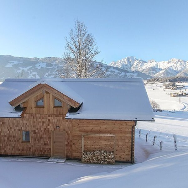 Der Hof in Holzbauweise mit schneebedecktem Dach und einem Holzstapel davor, gelegen in einer verschneiten Landschaft mit Bergpanorama.