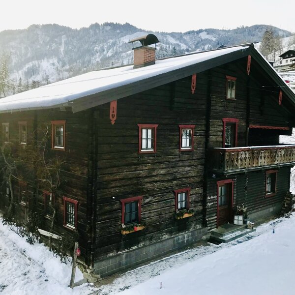 Der traditionelle Hof mit dunkler Holzfassade, Balkon und überdachtem Sitzbereich, umgeben von Schnee und Bergen.