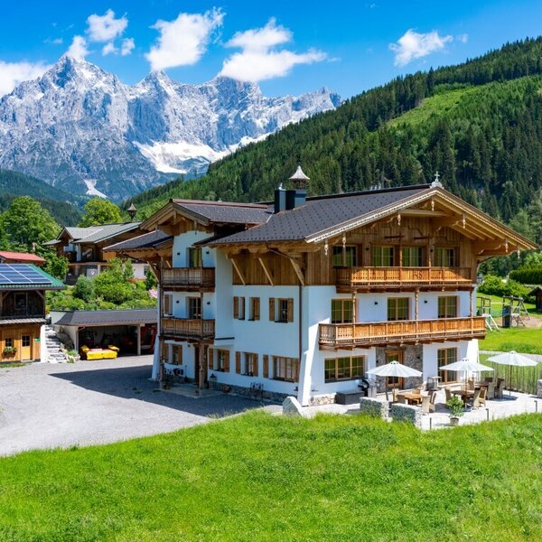 The farm house, featuring traditional wooden balconies and an outdoor patio with seating, is nestled in a green valley with a backdrop of forested hills and snowy mountain peaks.