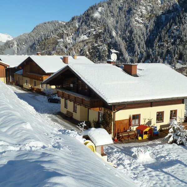 Der Bergbauernhof im Winter mit schneebedeckten Dächern, umgeben von verschneiter Berglandschaft und ausgestattet mit Spielgeräten im Garten.