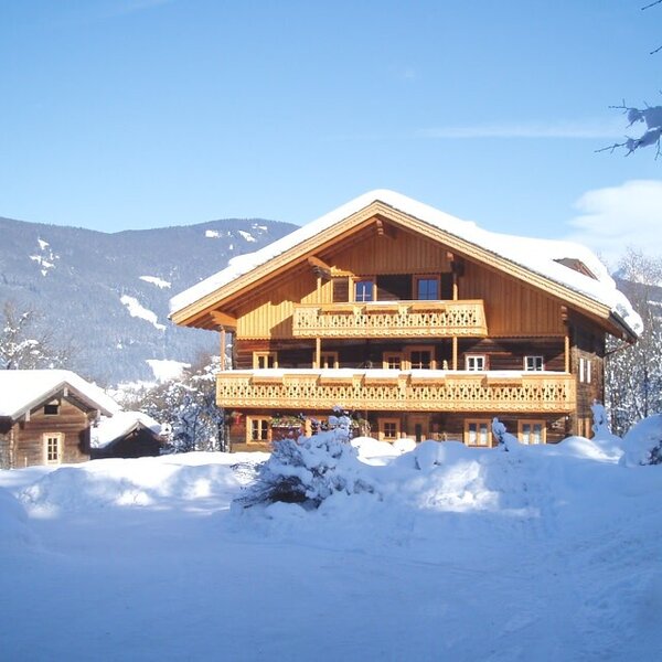 The multi-story wooden farm house, featuring carved balconies and a snow-covered roof, set against a winter mountain landscape.