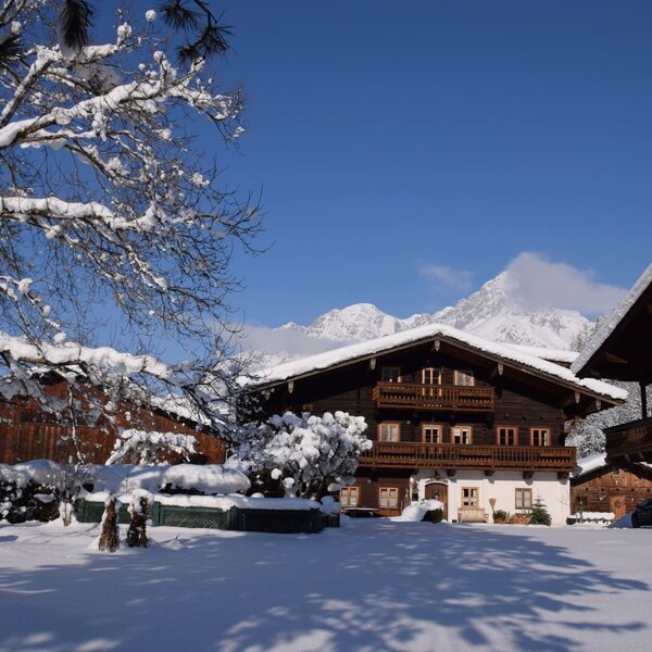 The Kendlhof farmhouse exterior in winter, featuring snow-covered traditional buildings and grounds set against snow-capped mountains.